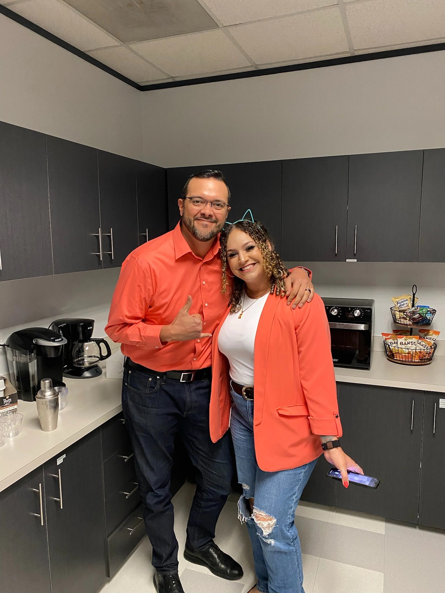 Man and woman in orange outfits posing in an office kitchen with coffee maker and snacks.