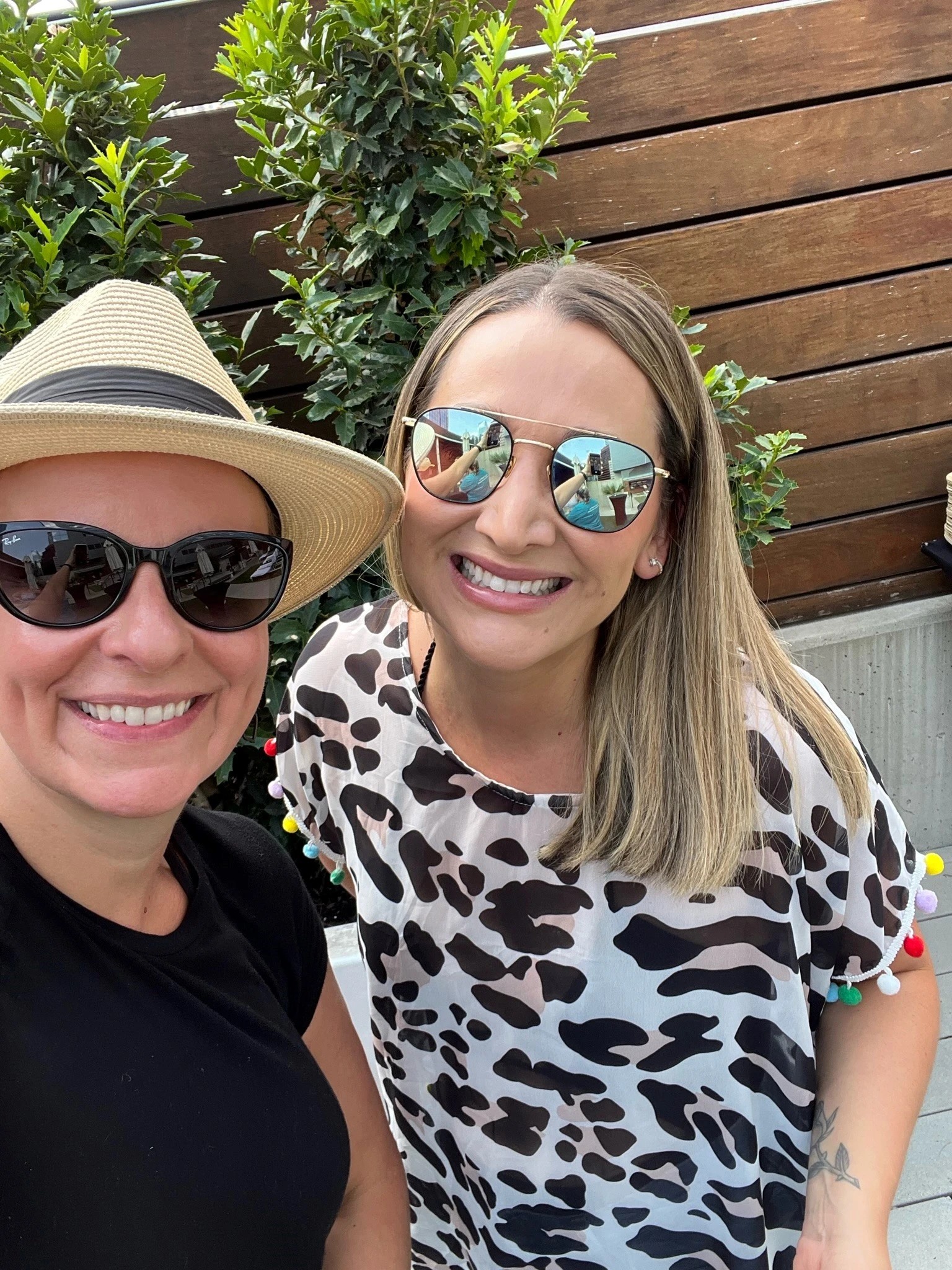 Two smiling women wearing sunglasses and hats take a selfie outdoors with lush greenery and wooden fencing behind them.