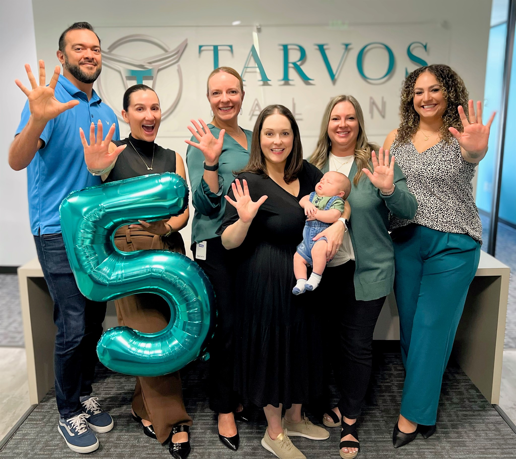 Group of people celebrating, holding a large "5" balloon. One person holds a baby.