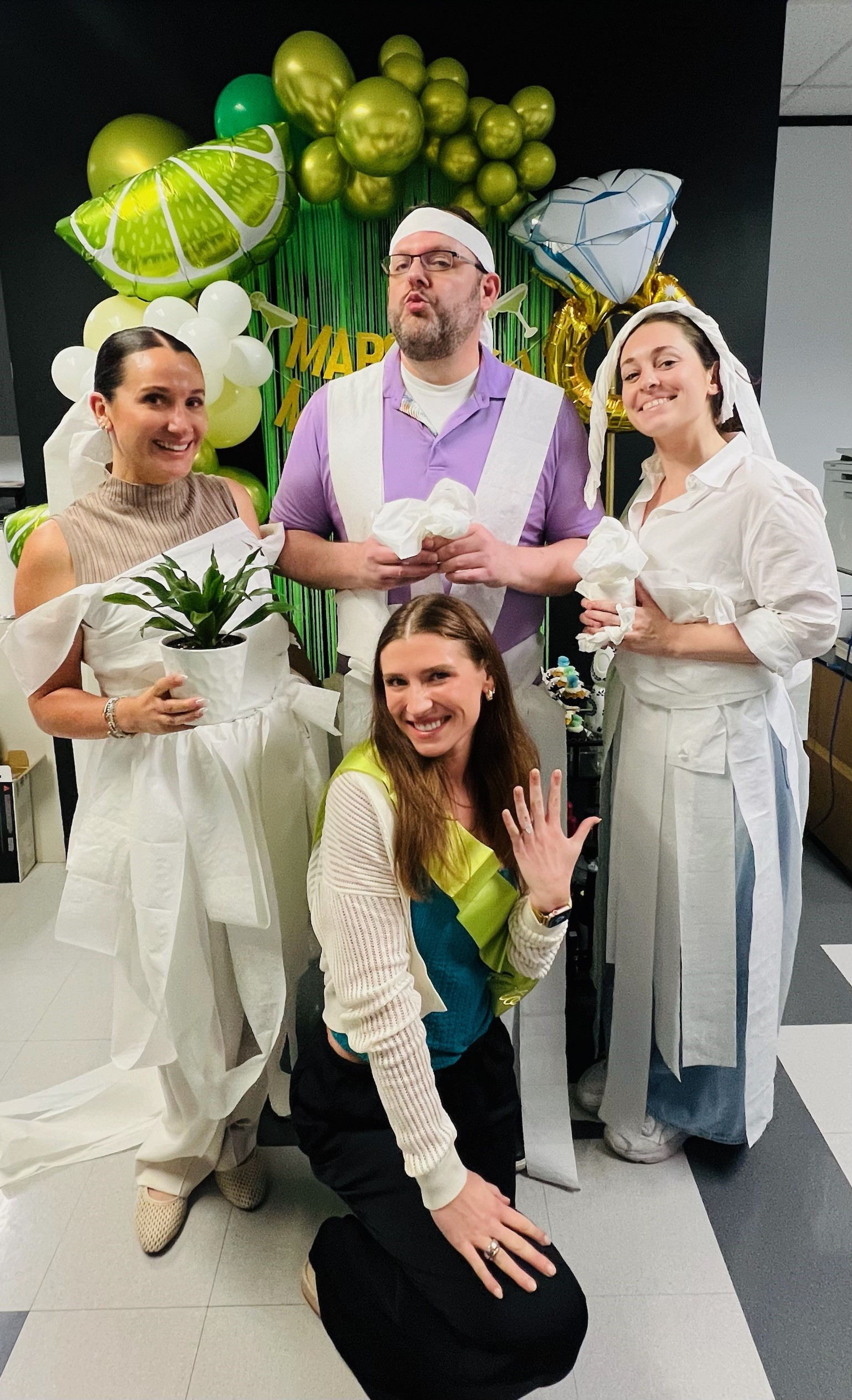 Friends in toilet paper wedding dresses and groom outfits at a party with lime and diamond balloons.