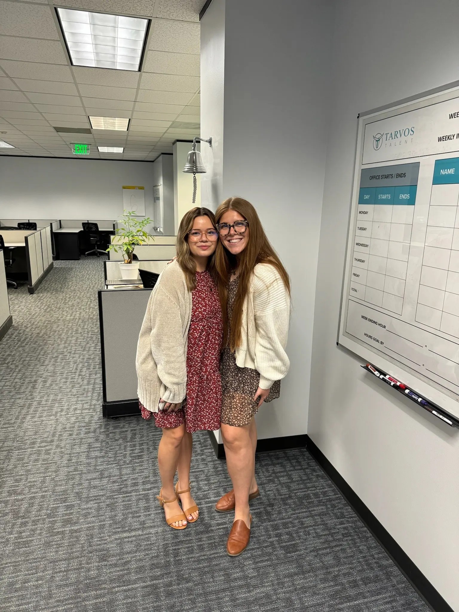 Two women in dresses and cardigans stand next to a whiteboard in a modern office setting.
