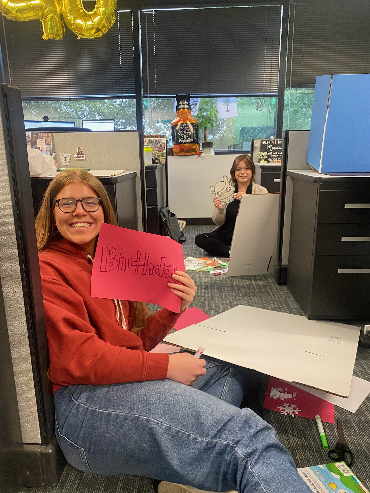 Smiling woman holds pink sign with "Birthday" written in black marker in an office.